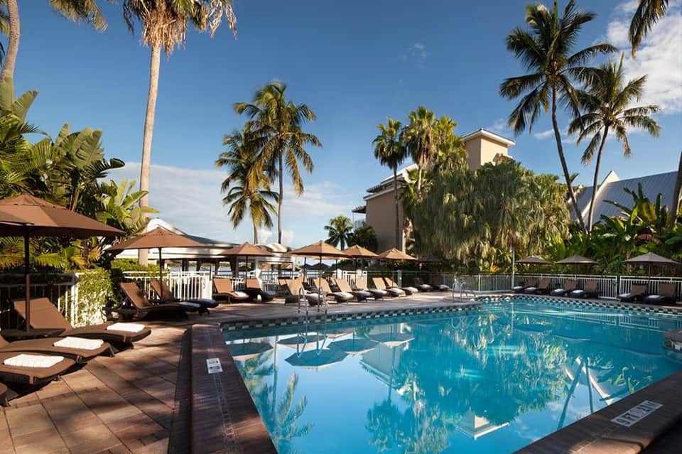 Resort pool surrounded by palm trees, lounge chairs, and tan umbrellas with multi-story hotel building