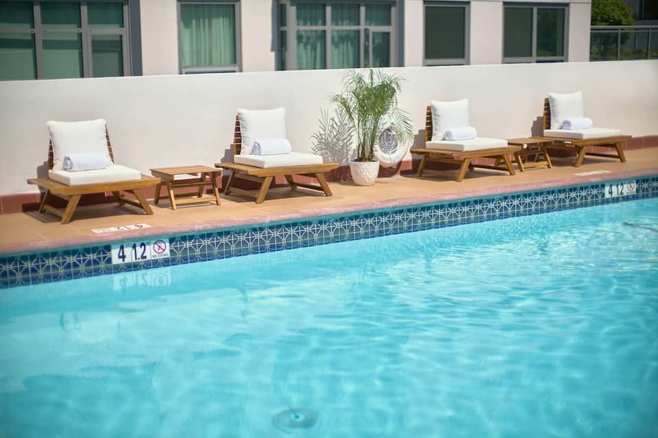 Outdoor pool area with wooden lounge chairs, white cushions, green plants, and modern white building facade