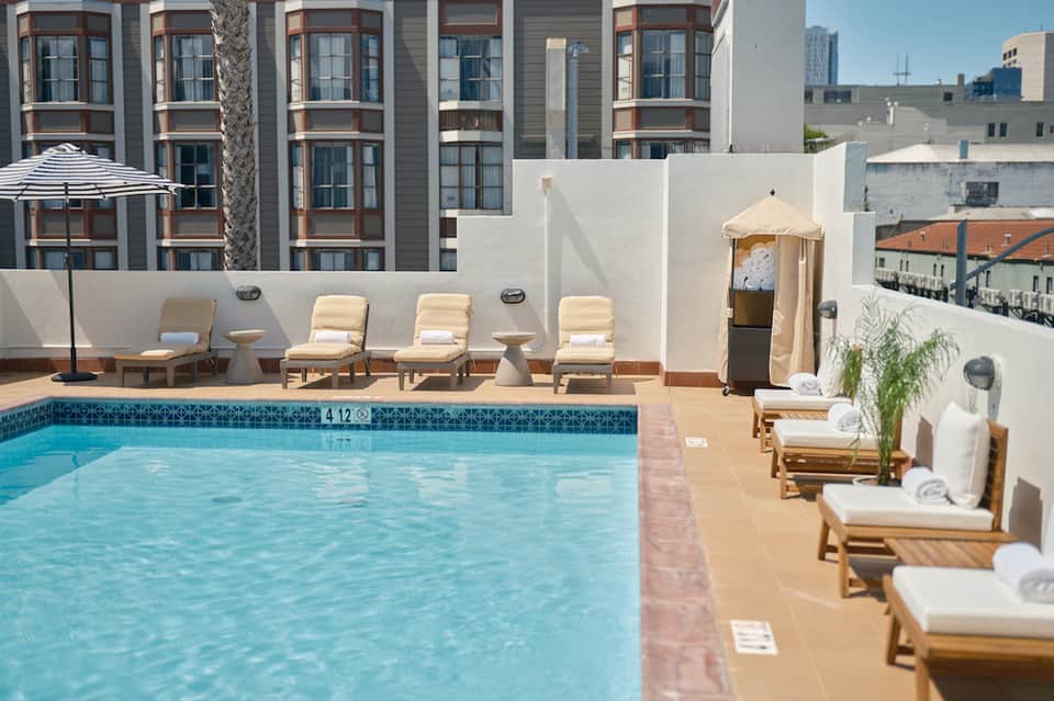 Outdoor rooftop pool with lounge chairs, umbrellas, and white wall backdrop facing tall residential building