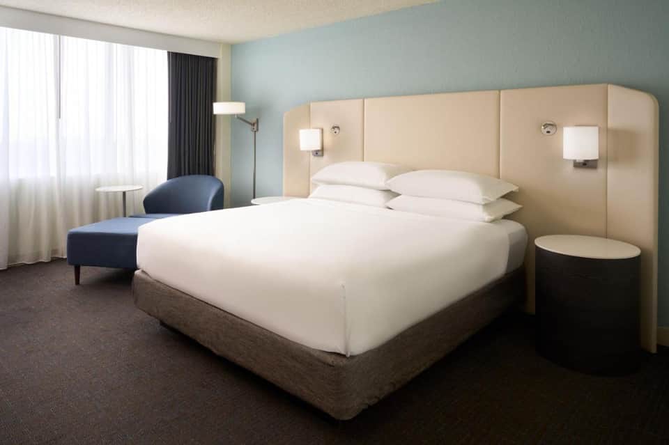Modern hotel bedroom with queen bed, beige headboard, light blue walls, and contemporary furnishings