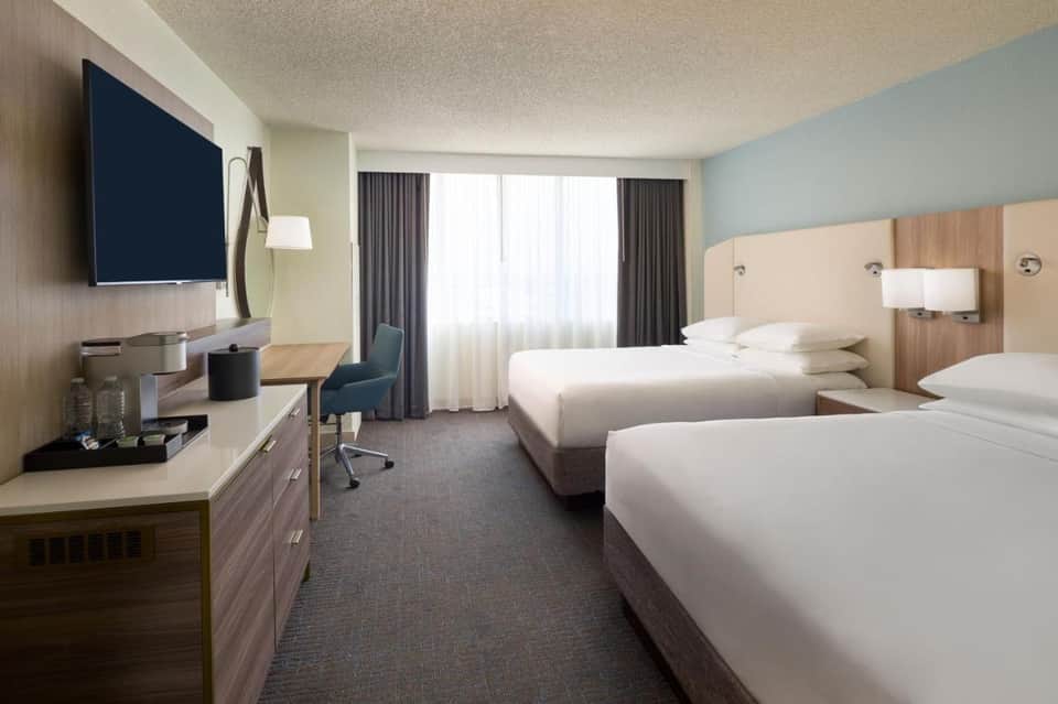 Modern hotel room with two queen beds, TV, desk, and large windows with curtains
