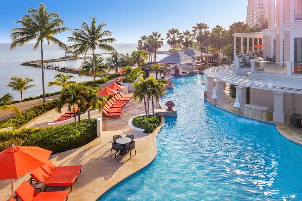 Luxury resort pool area with red loungers, palm trees, waterfront views, and modern architecture