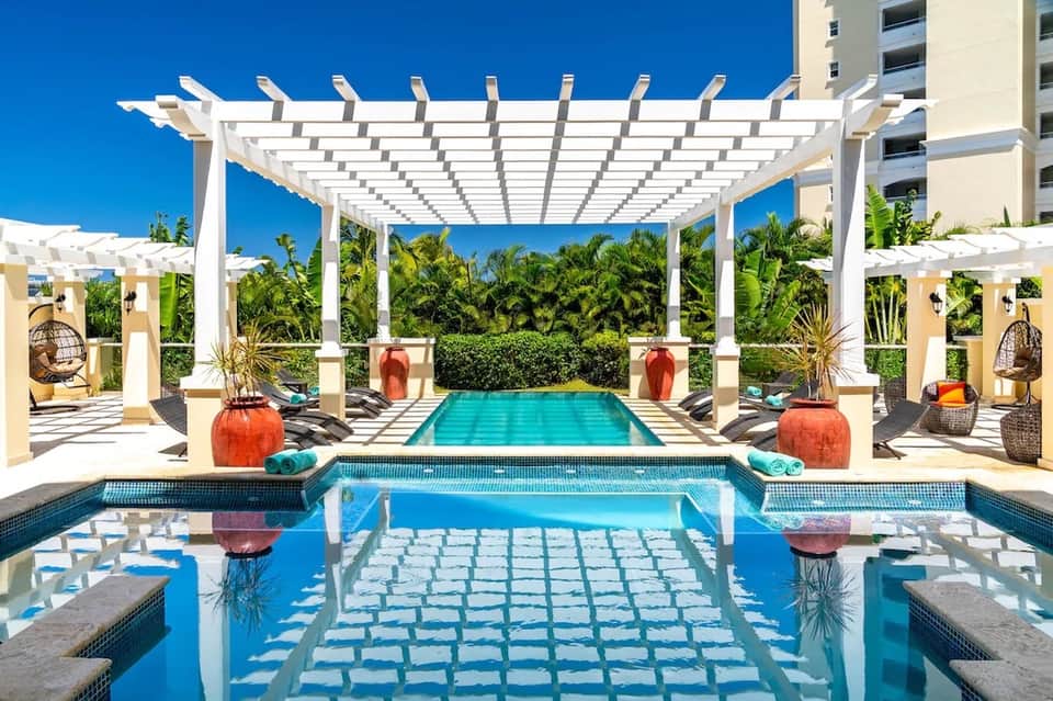 Resort pool with white pergola, spa jets, loungers, and lush tropical landscaping under blue sky