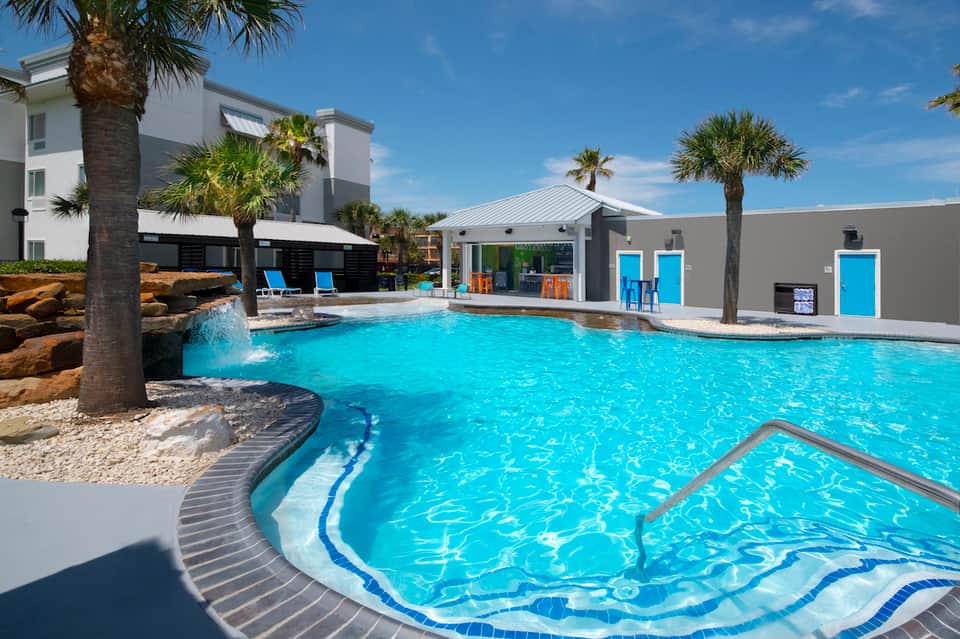 Resort pool area with turquoise water, palm trees, lounge chairs, and modern white pool house facility