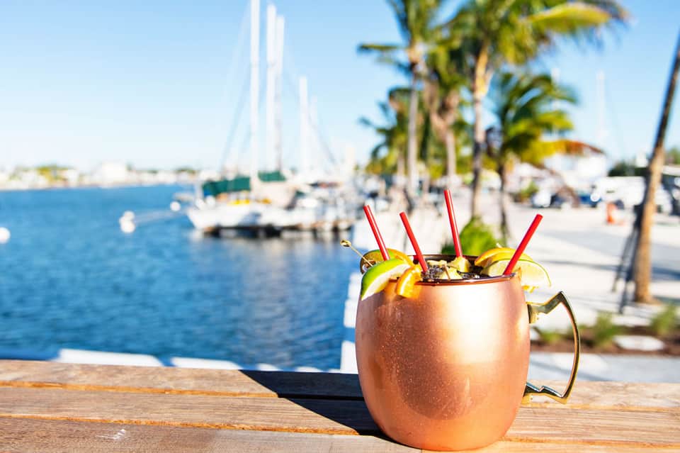 Tropical cocktail in copper mug with lime garnish on wooden dock, sailboats and palm trees in background.