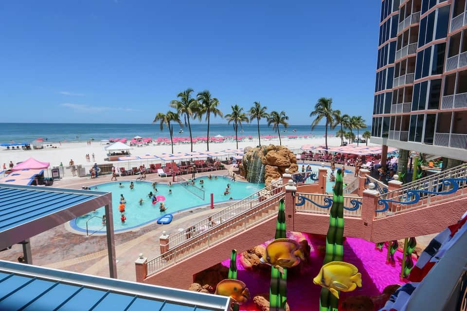 Beachfront resort pool with pink umbrellas, palm trees, ocean view, high-rise hotel building, and sandy beach