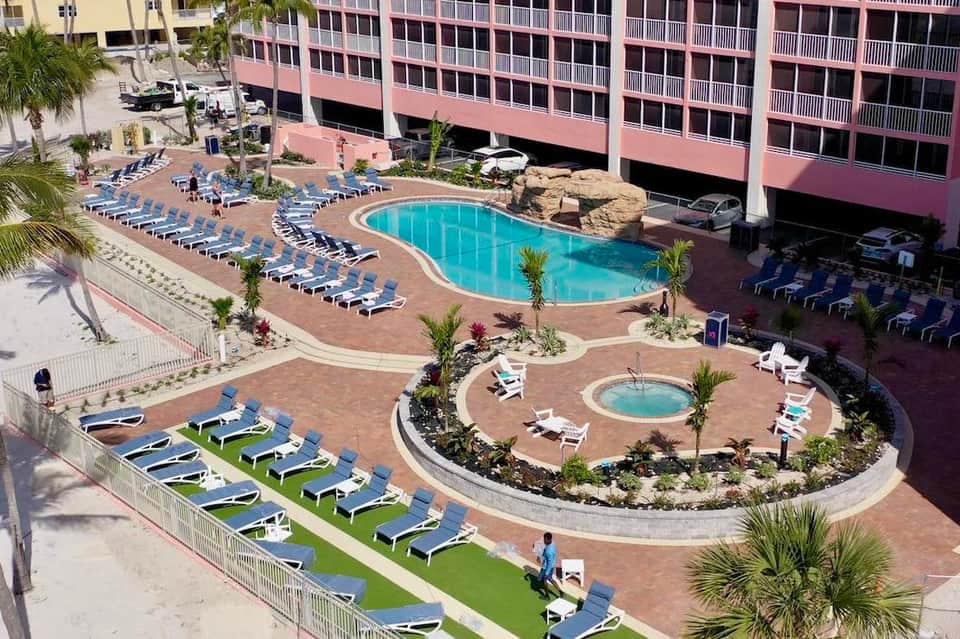 Aerial view of curved resort pool with lounge chairs, hot tub, and palm trees