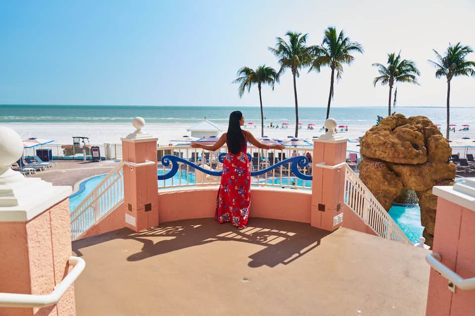 Woman in red floral dress stands on terrace railing overlooking beach with palm trees and ocean