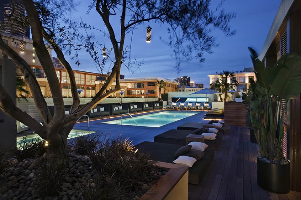 Modern rooftop pool at dusk with lounge beds, city skyline, and dramatic tree lighting