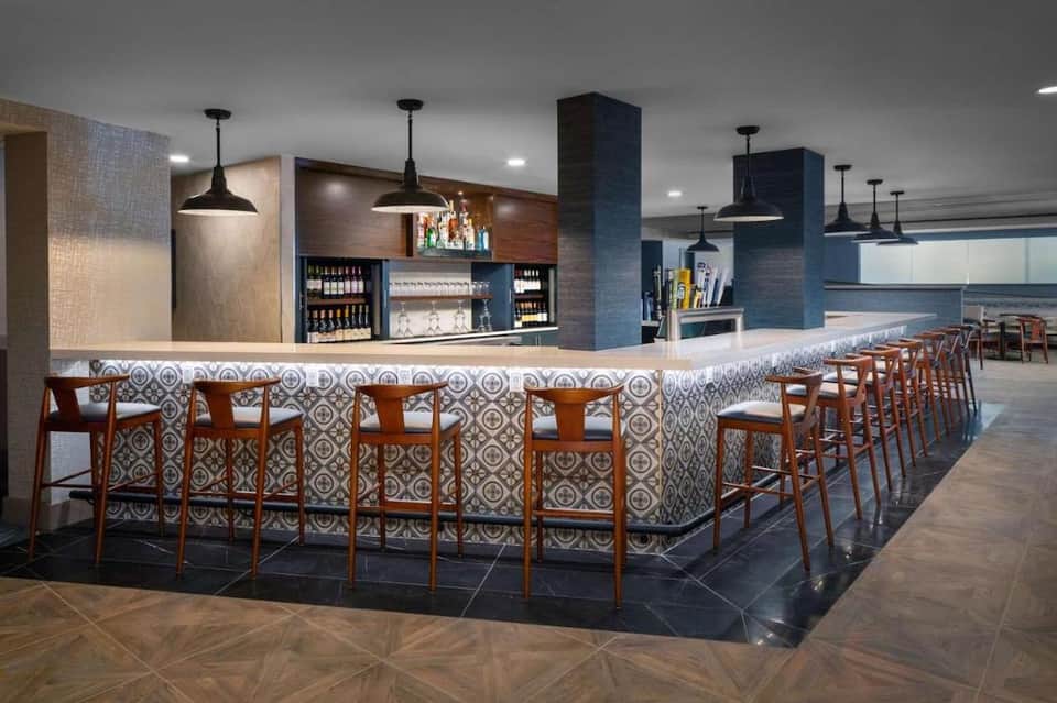 Contemporary hotel bar with patterned tile accents, wooden bar stools, pendant lighting, and shelved beverages