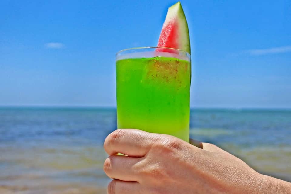 Hand holding green tropical cocktail with watermelon garnish against ocean beach background