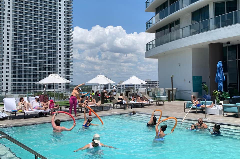 Rooftop pool deck with guests swimming and relaxing, white umbrellas, lounge chairs, and city skyline