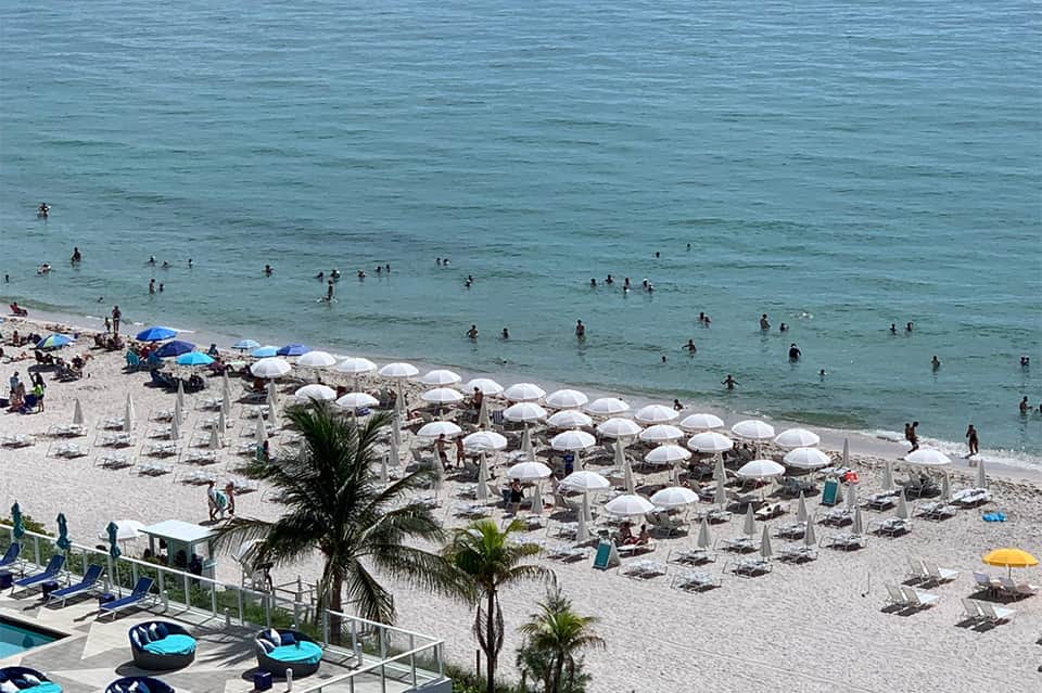 Beachfront with rows of white umbrellas, lounge chairs, swimmers in turquoise ocean, and palm trees