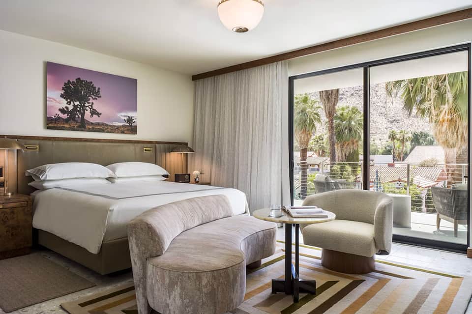 Luxurious hotel bedroom with king bed, artwork, modern furnishings, and balcony with palm trees