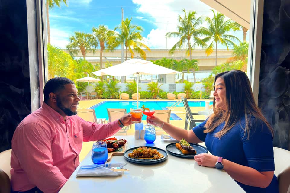 Couple toasting drinks at outdoor dining table overlooking resort pool with palm trees and tropical landscape.