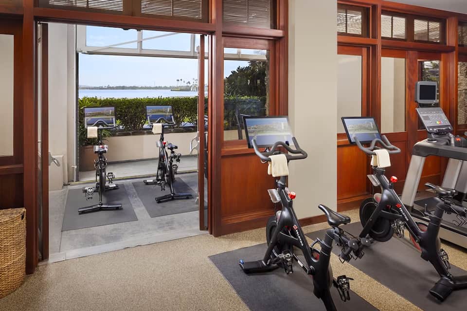 Indoor fitness studio with stationary bikes facing large windows overlooking waterfront and water activity equipment on patio