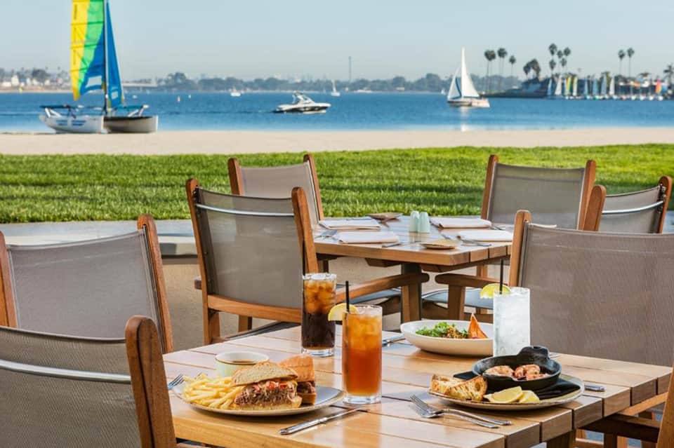 Waterfront dining area with wooden tables, sailboats visible on blue water, and palm trees in background