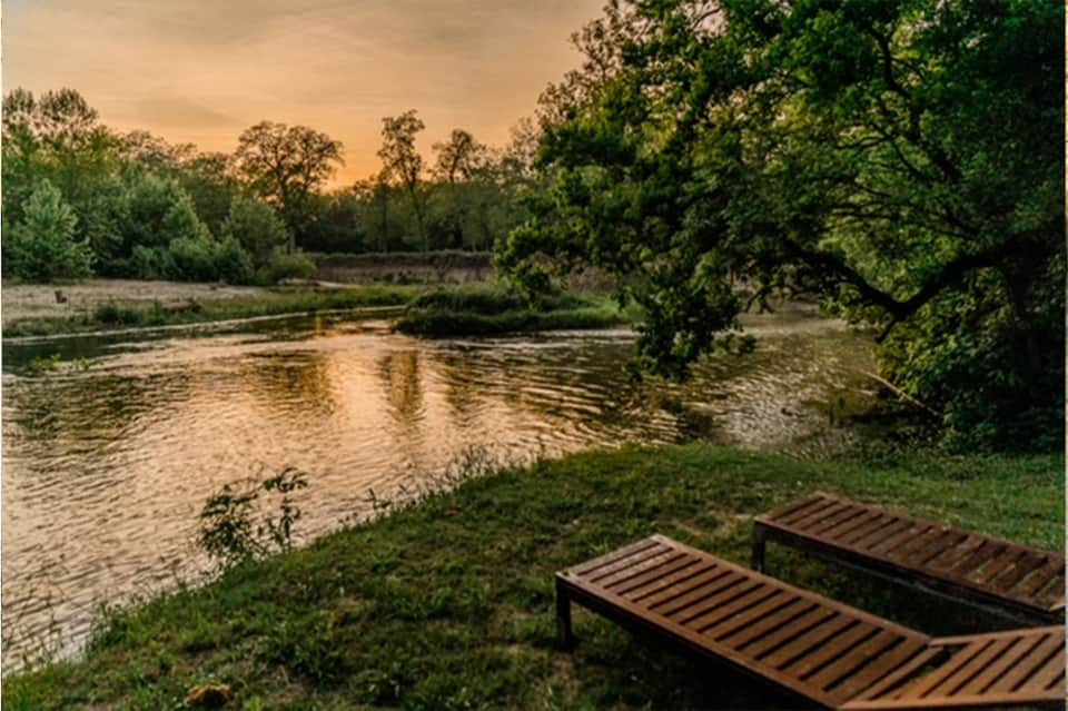 Wooden lounge chairs on grass overlooking a river at golden hour with trees