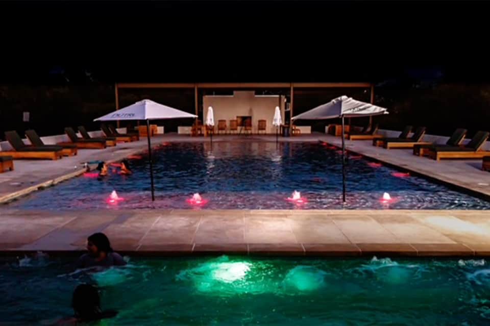 Nighttime resort pool with illuminated water features, lounge chairs under umbrellas, and ambient lighting