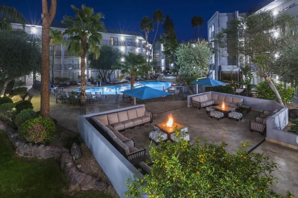 Resort courtyard at night with illuminated pool, fire pits with seating, palm trees, and modern white buildings
