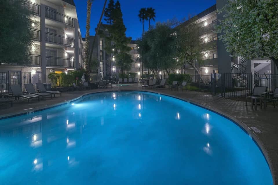 Illuminated resort pool at dusk surrounded by multi-story buildings with tropical landscaping