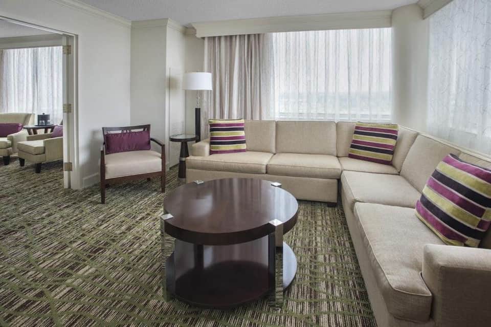 Spacious hotel suite with beige sectional sofa, round coffee table, and city views