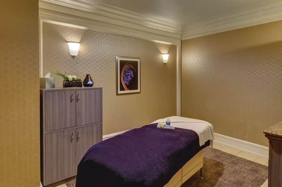 Spa treatment room with massage table, purple towel, wooden cabinetry, and warm ambient lighting
