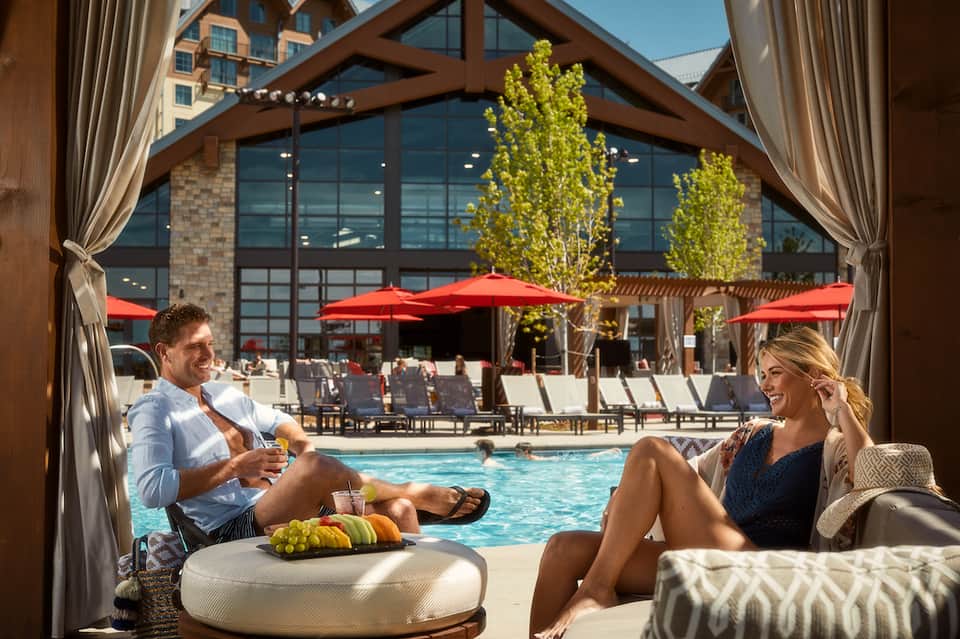 Couple relaxing on lounge chairs by pool under wooden pergola with red umbrellas and resort building