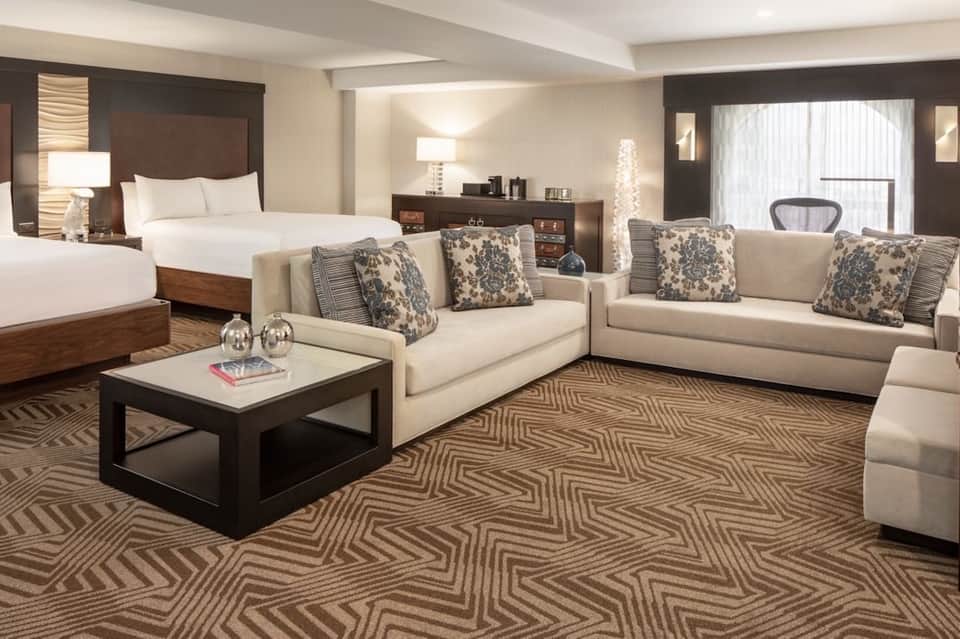Luxury hotel suite with king bed, cream sofas, patterned pillows, and dark wood accents