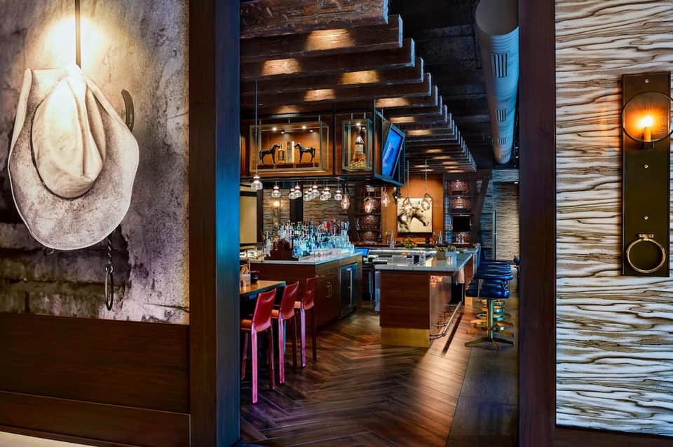 Dimly lit upscale bar entrance with cowboy hat artwork, dark wood accents, illuminated shelving, and reddish bar stools