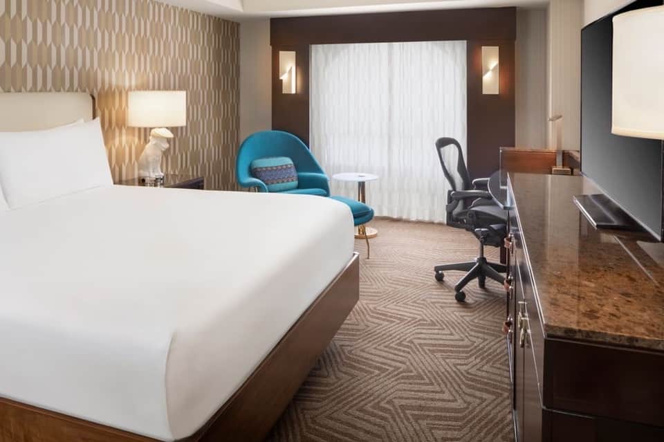 Modern hotel bedroom with king bed, blue accent chair, work desk, and partial view of bathroom