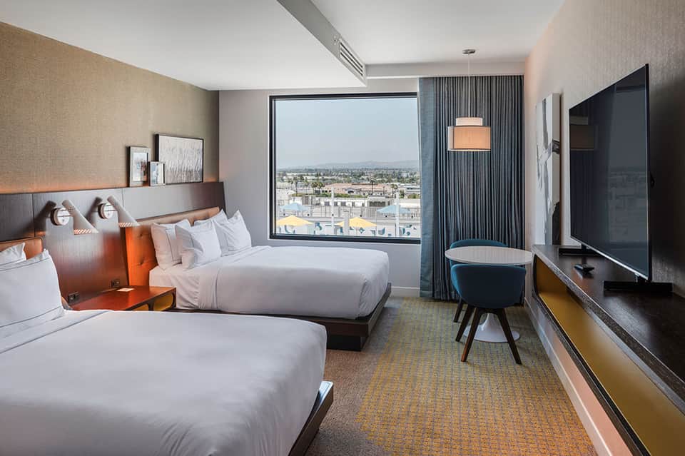Modern hotel room with two queen beds, city skyline view through floor-to-ceiling window