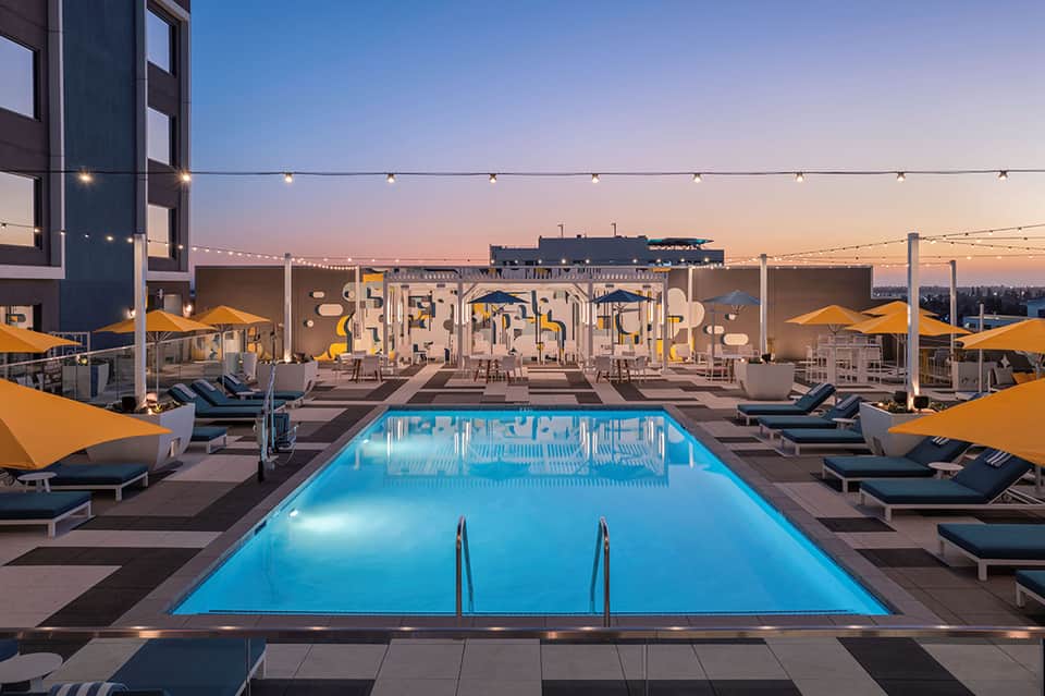 Rooftop pool at dusk with blue water, lounge chairs, yellow umbrellas, and cityscape with lit buildings