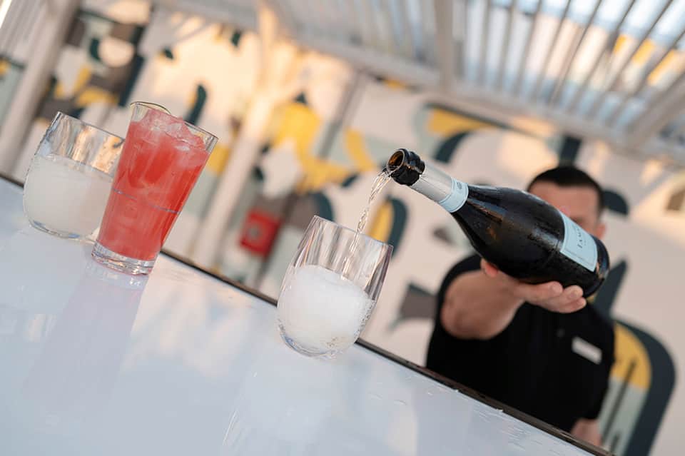 Champagne bottle pouring into glass with red and clear beverages on outdoor patio