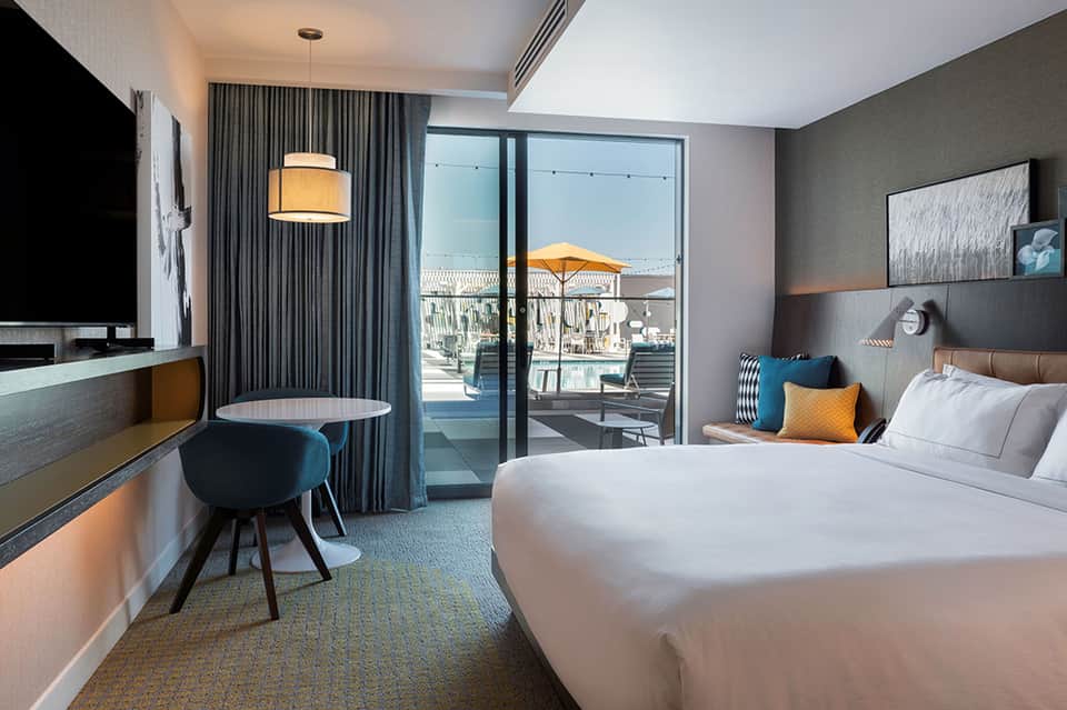 Modern hotel bedroom with king bed, desk, flat-screen TV, and sliding glass door to ocean-view balcony
