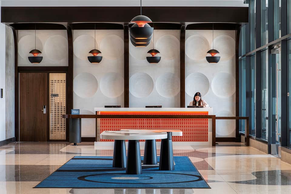 Modern hotel lobby reception desk with orange accent wall, decorative pendant lights, and geometric backdrop