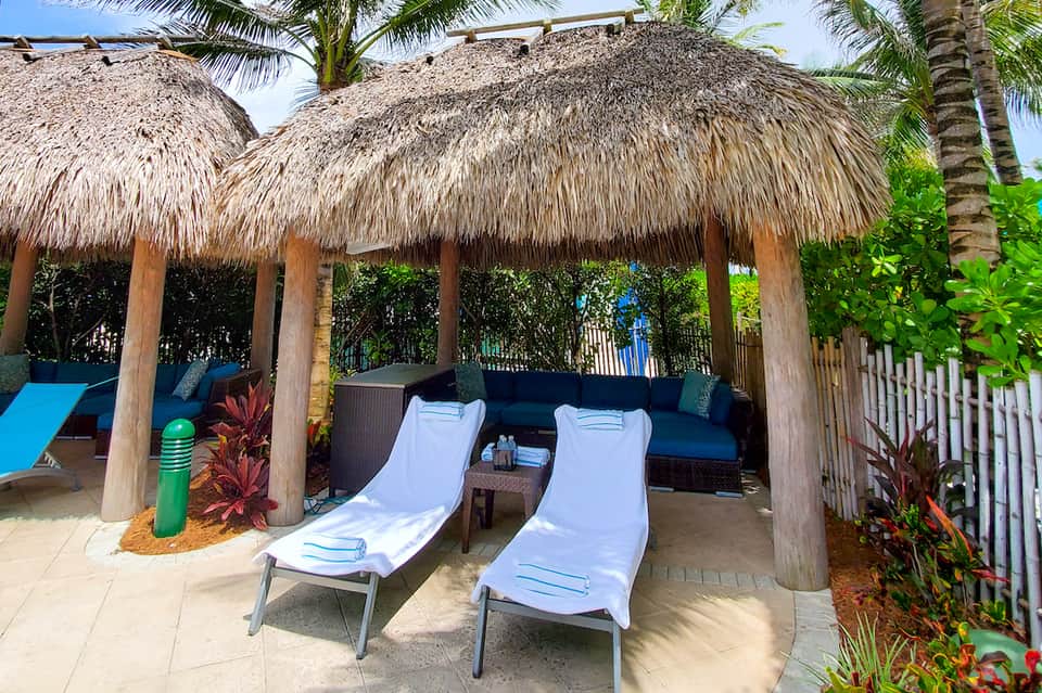Beachside cabana with thatched roof covering lounge chairs and wicker seating near palm trees