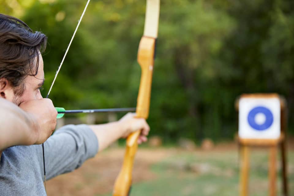 Man practicing archery outdoors with a wooden bow and green arrow at a target