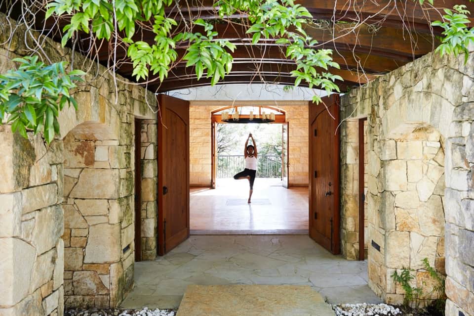 Woman in yoga pose on wooden platform visible through stone archway with green foliage overhead