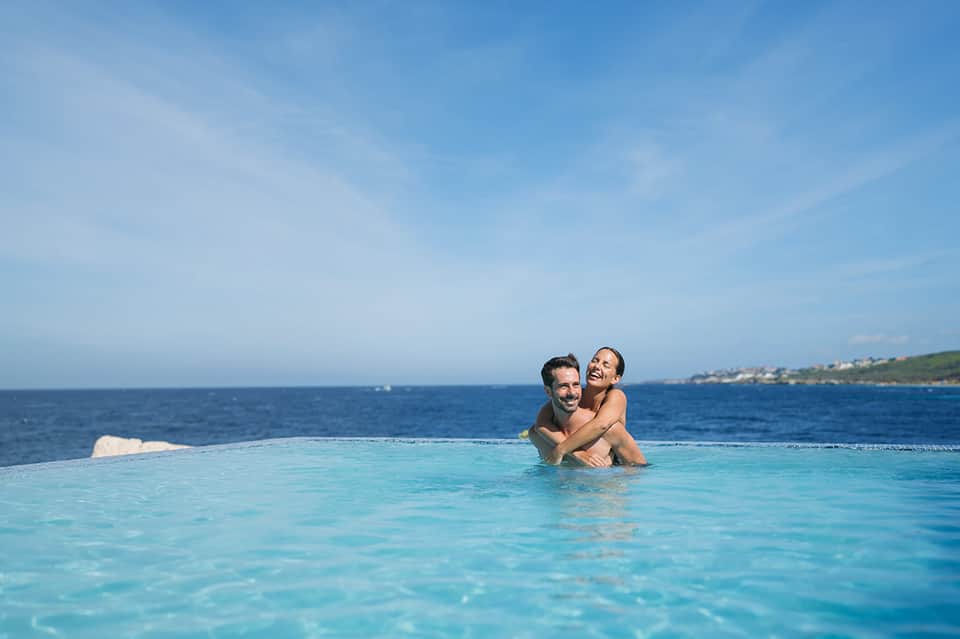 Couple enjoying infinity pool overlooking turquoise ocean and coastal landscape