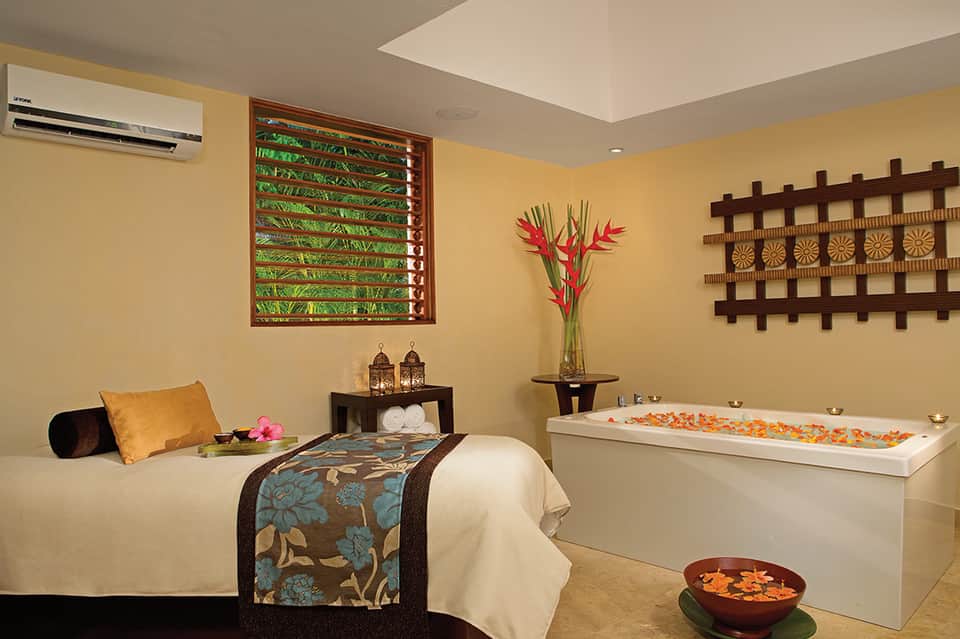 Luxurious spa room with soaking tub, massage bed, decorative flowers, and tropical-themed décor in warm gold tones