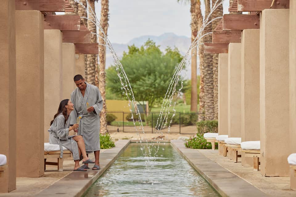 Couple in spa robes relaxing beside water feature with fountains and palm trees