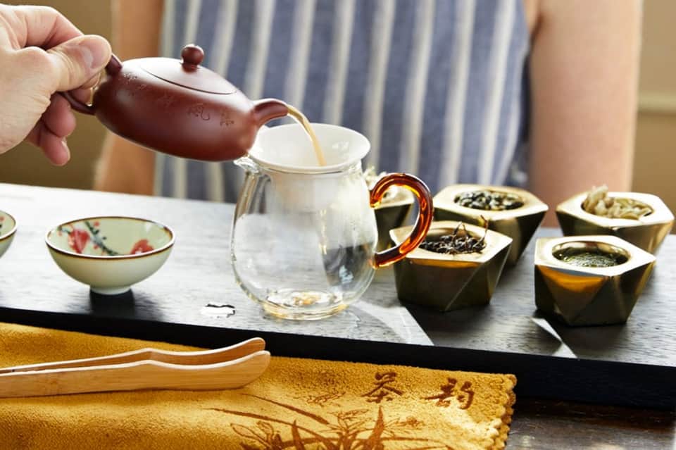 Tea ceremony setup with dark teapot pouring into glass cup and loose tea samples in bowls
