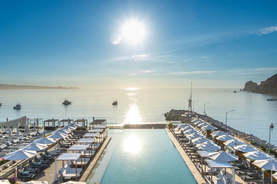 Oceanfront infinity pool with lounge chairs and blue umbrellas overlooking boats and coastal cliffs