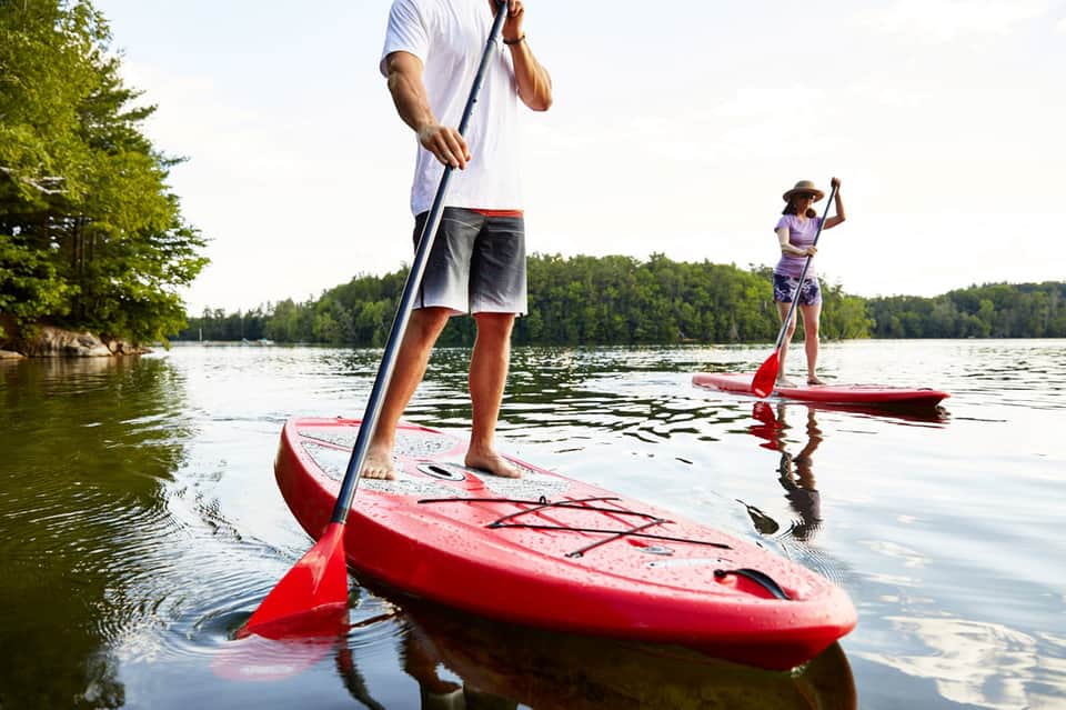 Two people paddleboarding on red kayaks in a calm lake surrounded by forested shoreline