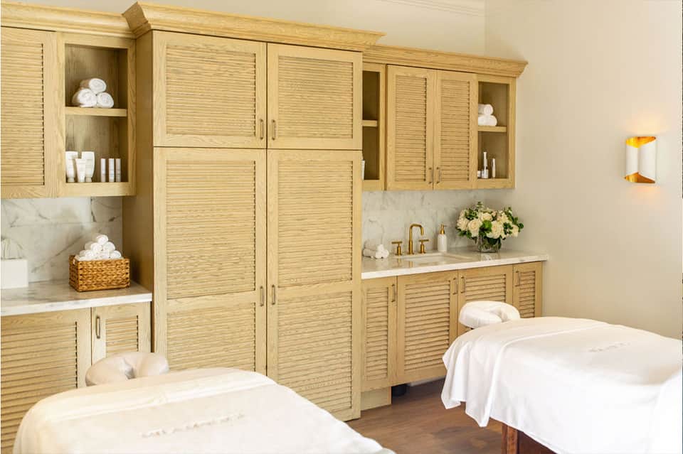 Spa treatment room with wooden cabinetry, marble countertops, two massage tables, and fresh flowers