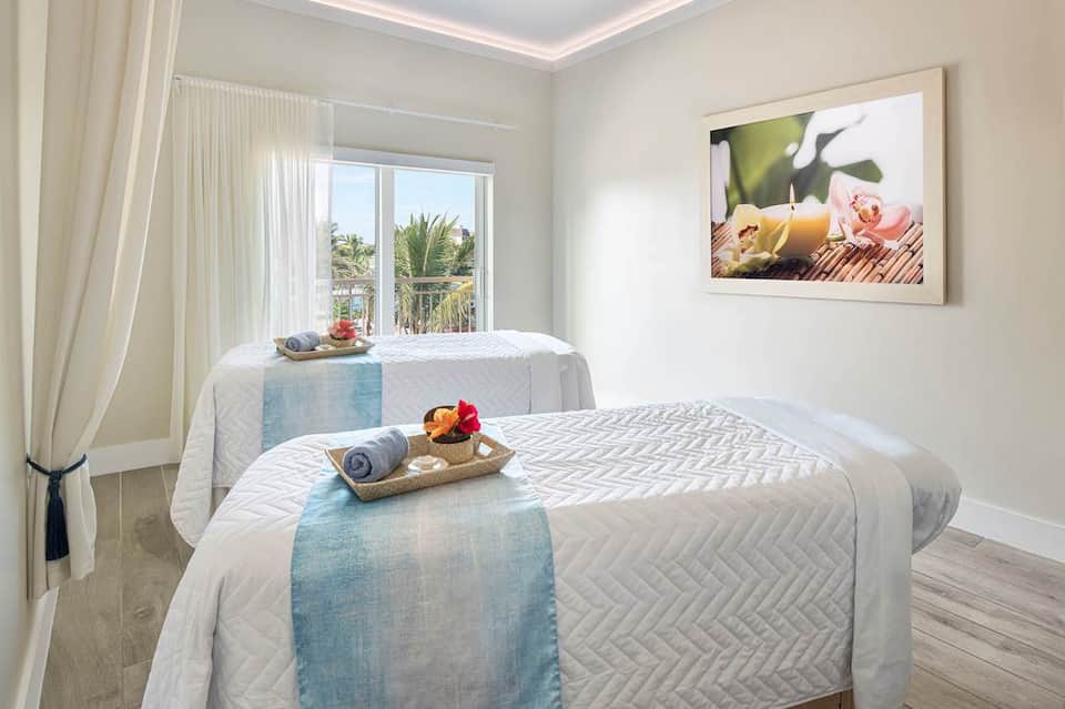 Spa treatment room with two massage tables, large windows overlooking palm trees, and calming décor