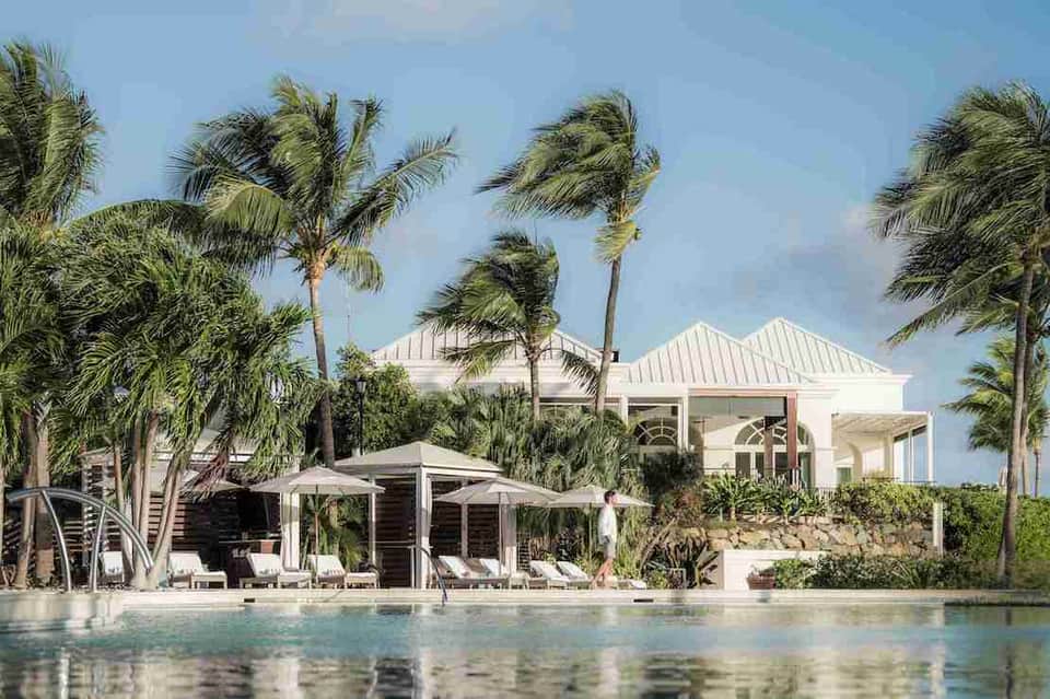 Waterfront villa with white architectural roofline, palm trees, lounge area, and pool overlooking water