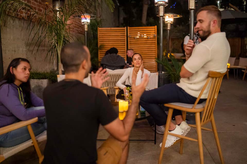 Group of people gathered outdoors at night on wooden chairs in casual conversation setting
