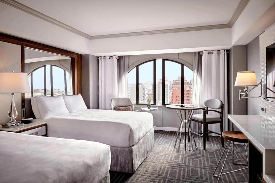 Modern hotel bedroom with two beds, city views through arched windows, and contemporary furnishings